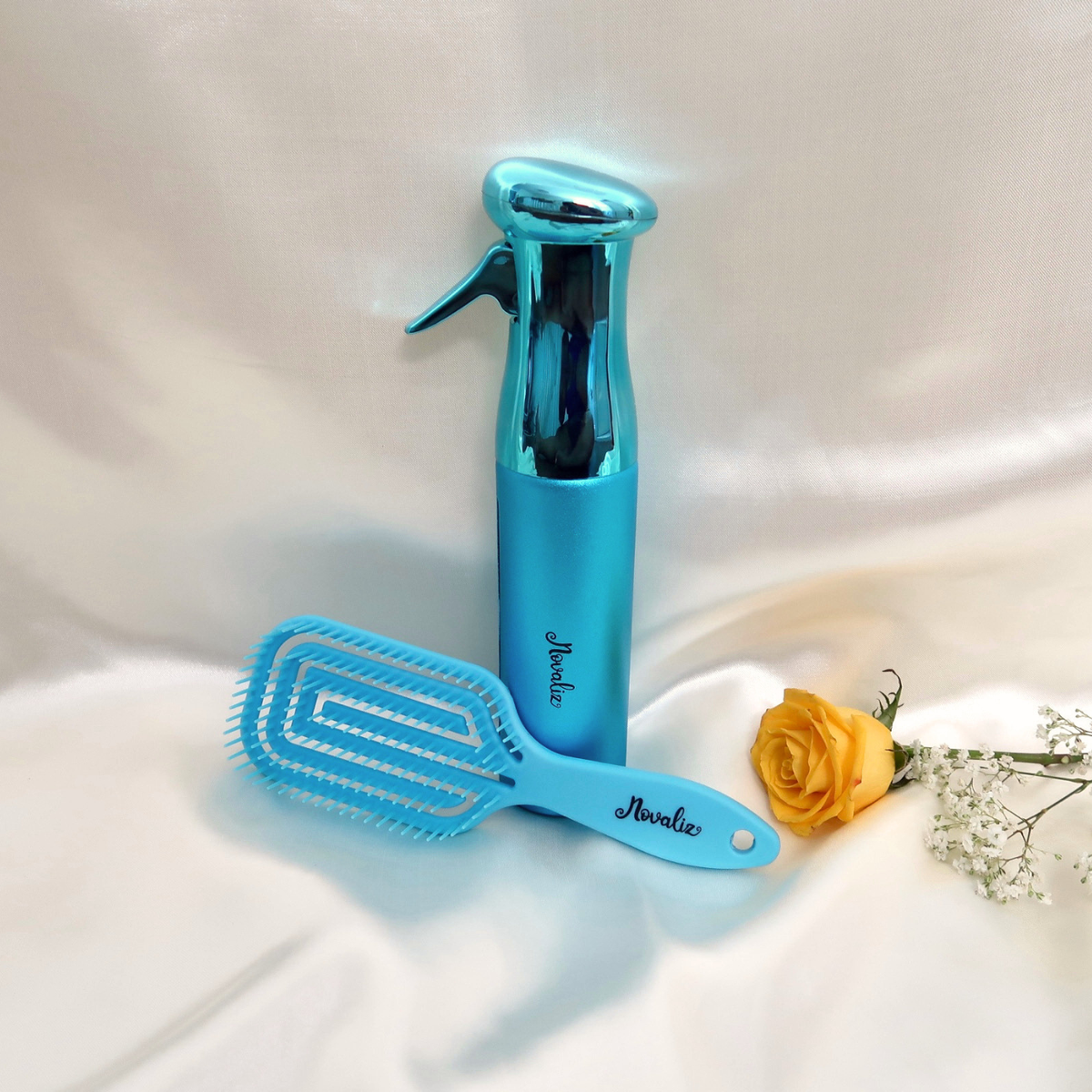 A blue flexible detangling hair brush styling set featuring a continuous mist spray bottle  both branded 'Novaliz', arranged on a draped white fabric background with a yellow rose and baby's breath prop.
