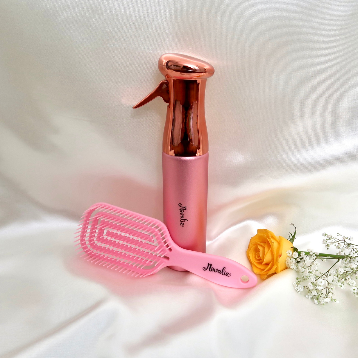 A pink flexible detangling hair brush styling set featuring a continuous mist spray bottle  both branded 'Novaliz', arranged on a draped white fabric background with a yellow rose and baby's breath prop.