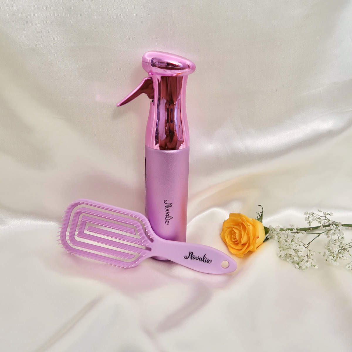 A lilac purple flexible detangling hair brush styling set featuring a continuous mist spray bottle both branded 'Novaliz', arranged on a draped white fabric background with a yellow rose and baby's breath prop.